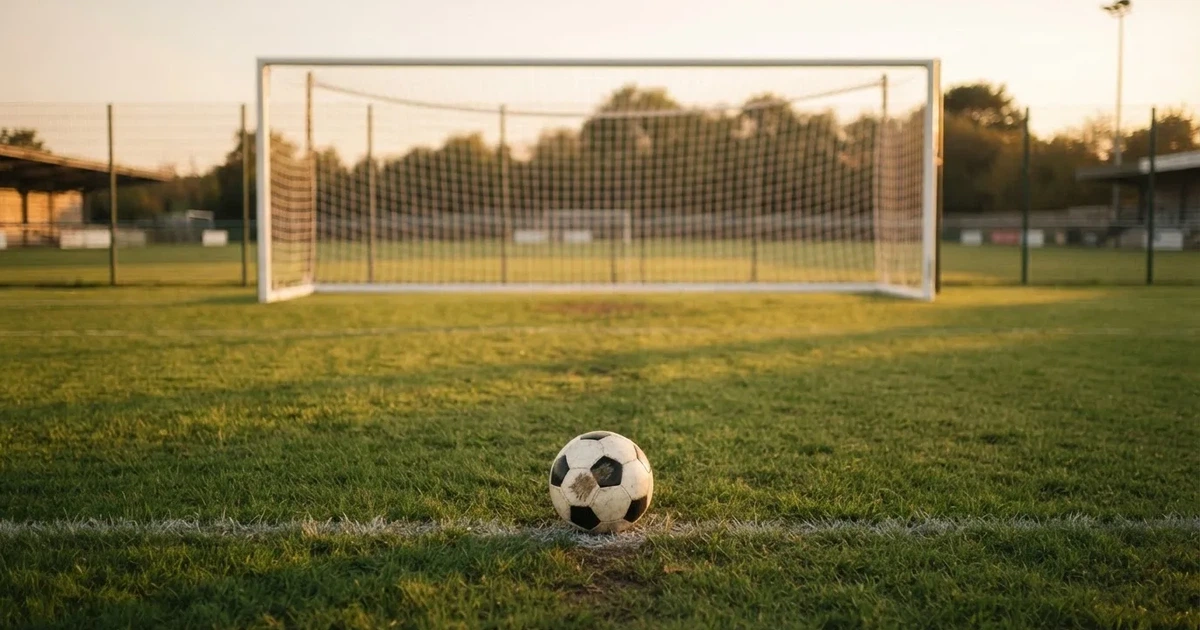Ein Fußball liegt vor einem leeren Tor auf dem Rasen, das Netz hängt unbewegt, im Hintergrund ein verschwommener Zaun in warmem Abendlicht.
