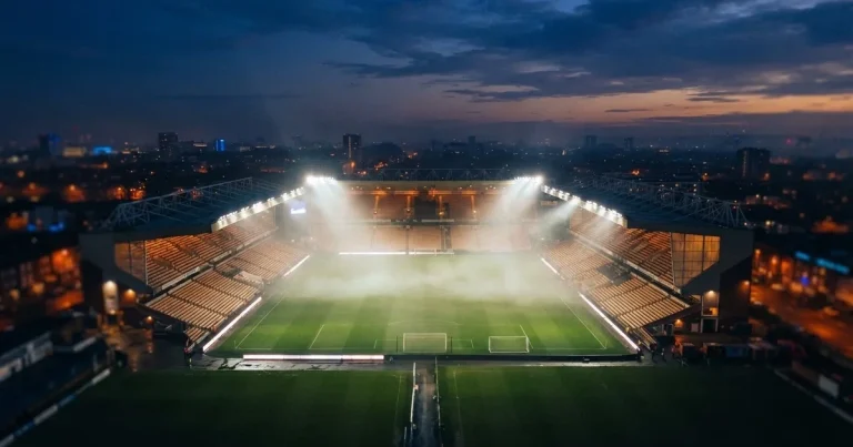 Ein Stadion unter Flutlicht aus der Ferne am Abend, hell erleuchtet, Nebel liegt über dem Rasen.