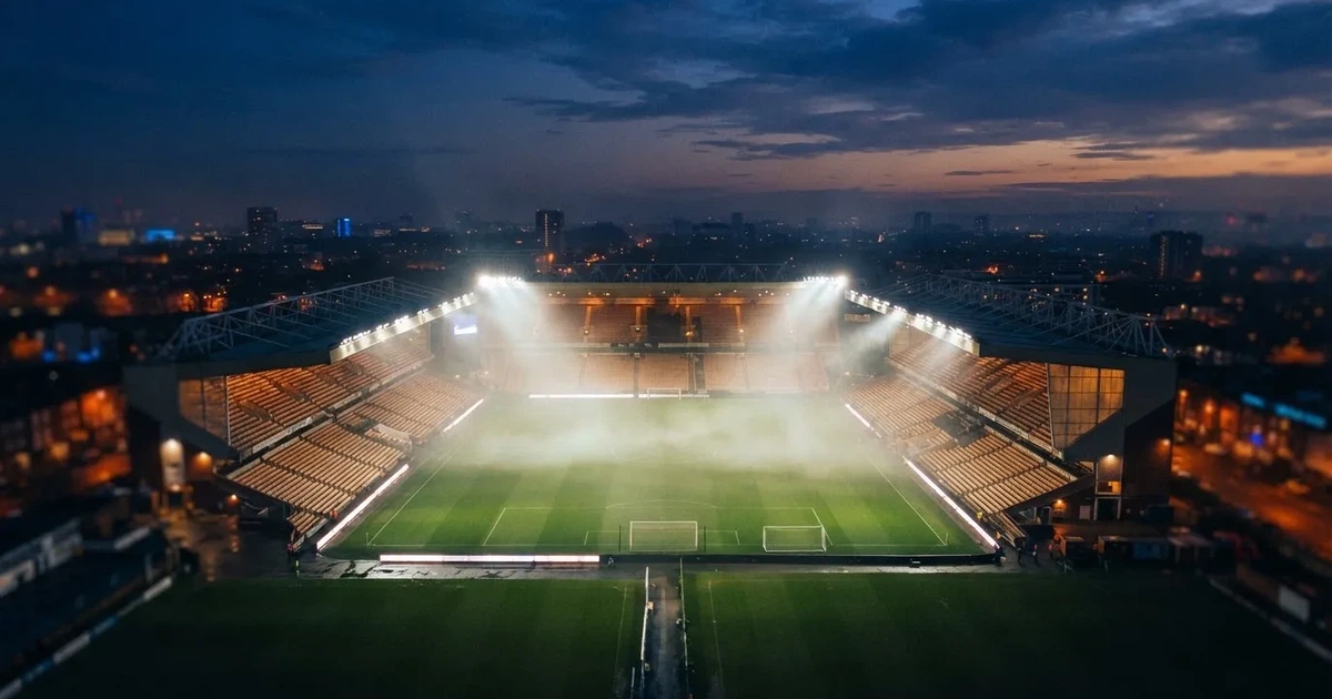 Ein Stadion unter Flutlicht aus der Ferne am Abend, hell erleuchtet, Nebel liegt über dem Rasen.