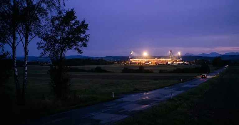 Ein Stadion unter Flutlicht aus großer Entfernung gesehen, davor eine dunkle Landstraße und einzelne Bäume in der Abenddämmerung.