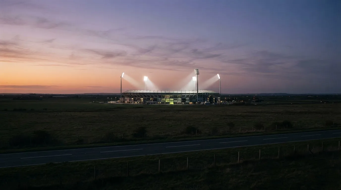 Ein Fußballstadion unter Flutlicht aus großer Entfernung gesehen, davor Felder und eine ruhige Straße in der Dämmerung.
