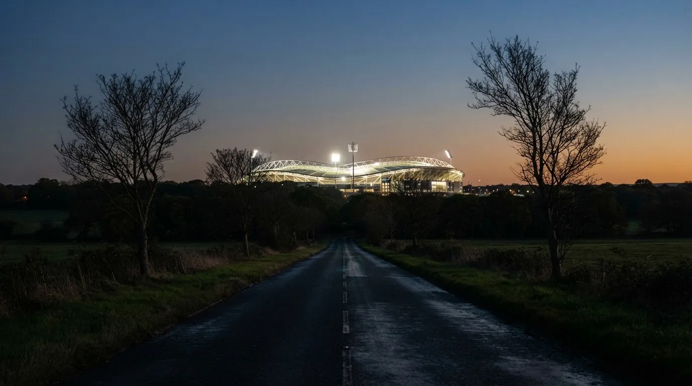 Ein Stadion unter Flutlicht aus großer Entfernung gesehen, davor eine dunkle Landstraße und einzelne Bäume in der Dämmerung.